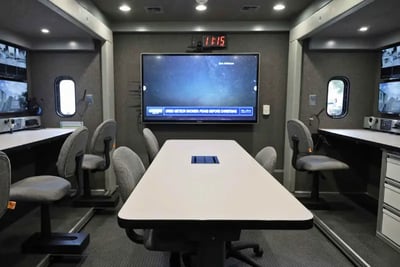 The interior of a mobile command vehicle showing a large monitor and conference table.