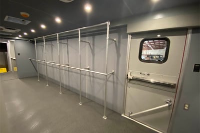 The inside of a mobile decontamination unit shows a large built-in storage rack system and an exit doorway with a window.
