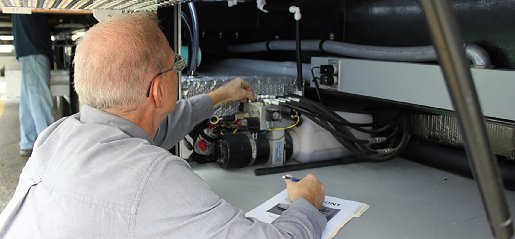 A man in a light-colored shirt works on the system mechanics of a specialty vehicle writing notes with a pen on paper.