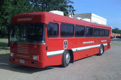 An old red city bus sitting on pavement in front of a building with blue sky and green trees in the background.