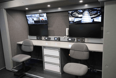 The interior of a Mobile Command Vehicle showing 4 screens above a desk with two chairs.