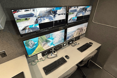 The interior of a Mobile Command Vehicle showing four screens mounted above a desk with two chairs and two keyboards sitting on it.