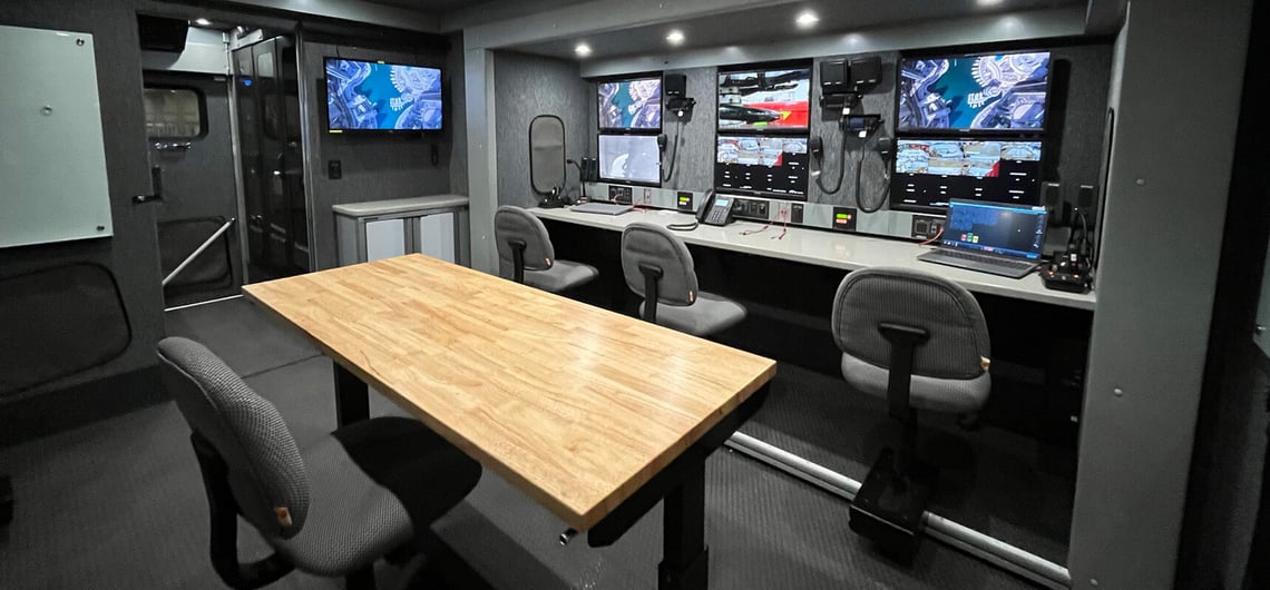 The inside of a hostage negotiation vehicle is black and gray with multiple screens, four chairs and a wood table in the center.