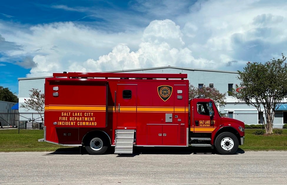 Frontline Communications red Mid-Size Mobile Command vehicle parked outside near the building on a cloudy day near trees. 