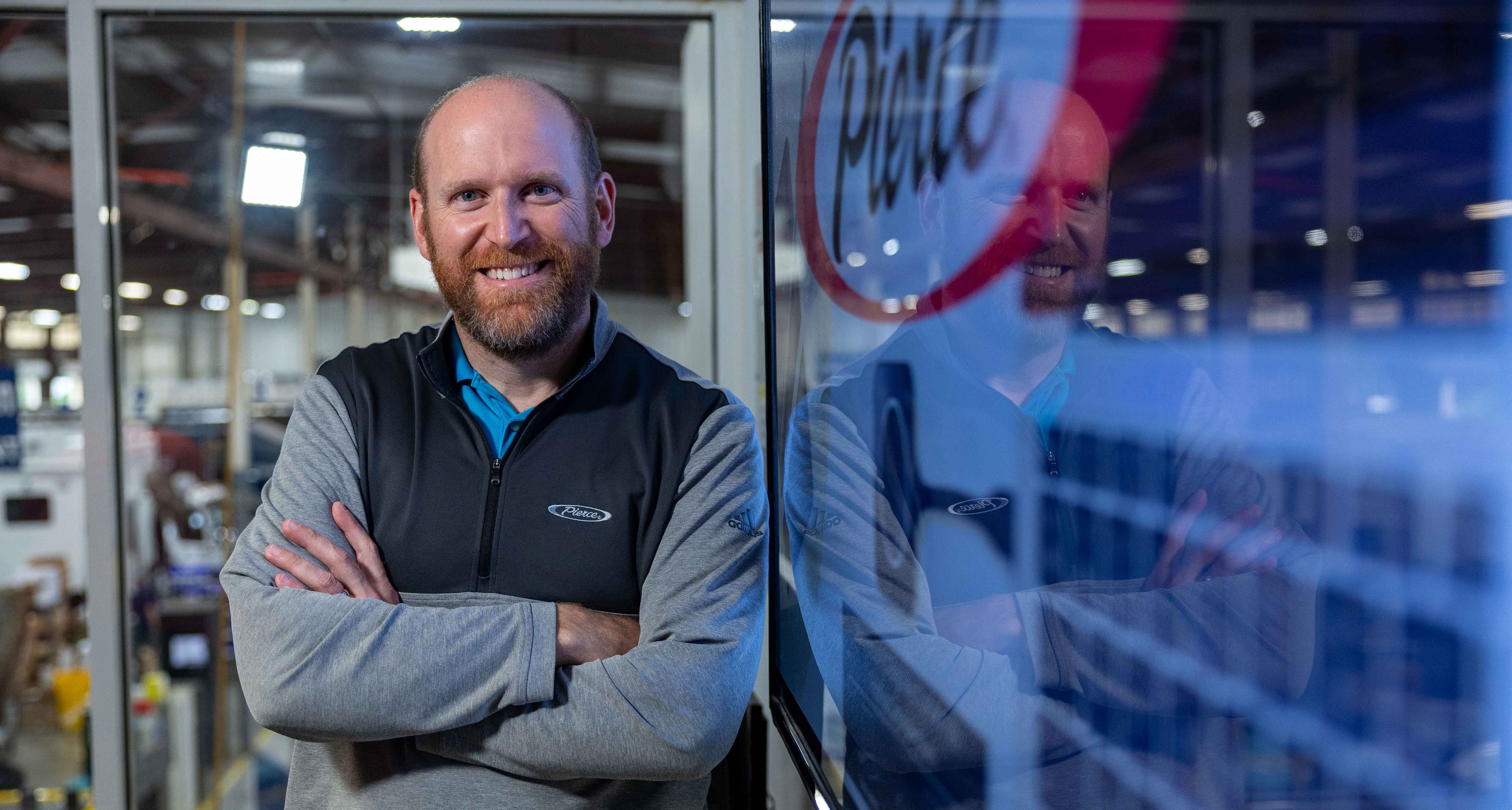 A man standing next to a television monitor displaying a Pierce logo with the Frontline Communications manufacturing facility behind him.